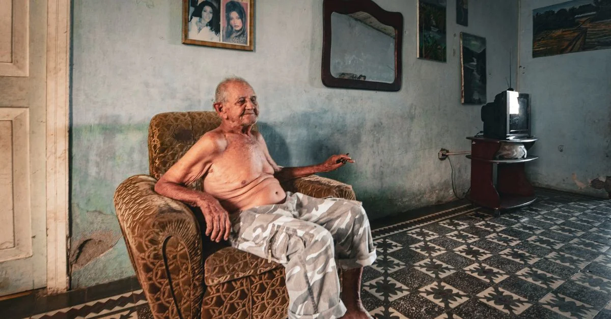 Un homme senior assis torse nu dans un fauteuil dans une pièce vintage, reflétant un moment paisible.