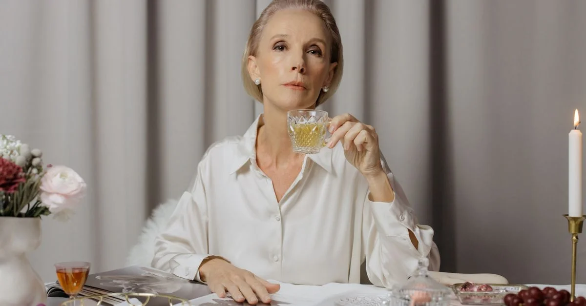 Femme senior en blouse blanche sirotant une boisson dans un cadre élégant avec des fleurs et des bougies.