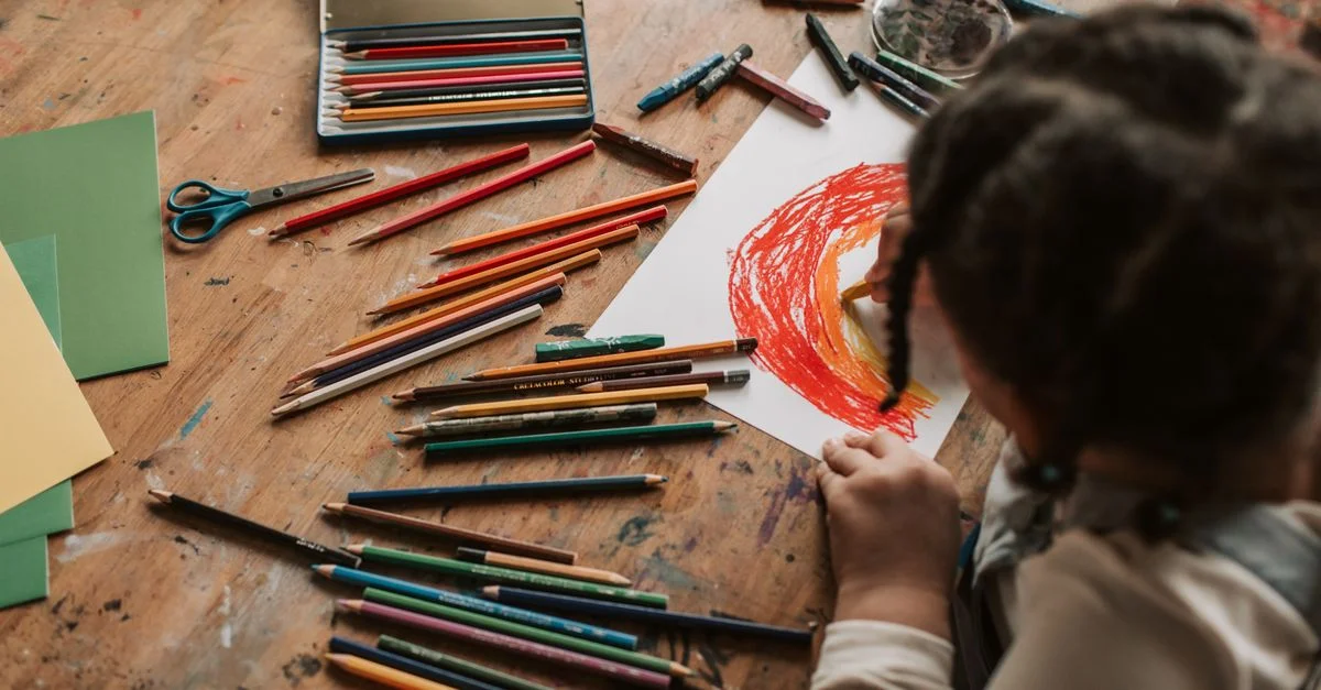 Une jeune enfant dessine une image colorée avec des crayons sur une table en bois, entourée de fournitures artistiques à l'intérieur.