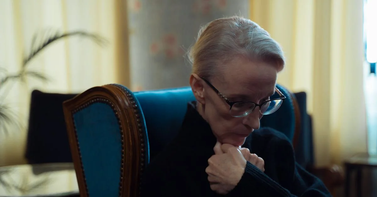 Une femme âgée portant des lunettes assise dans un fauteuil, perdue dans ses pensées dans une pièce chaleureusement éclairée.