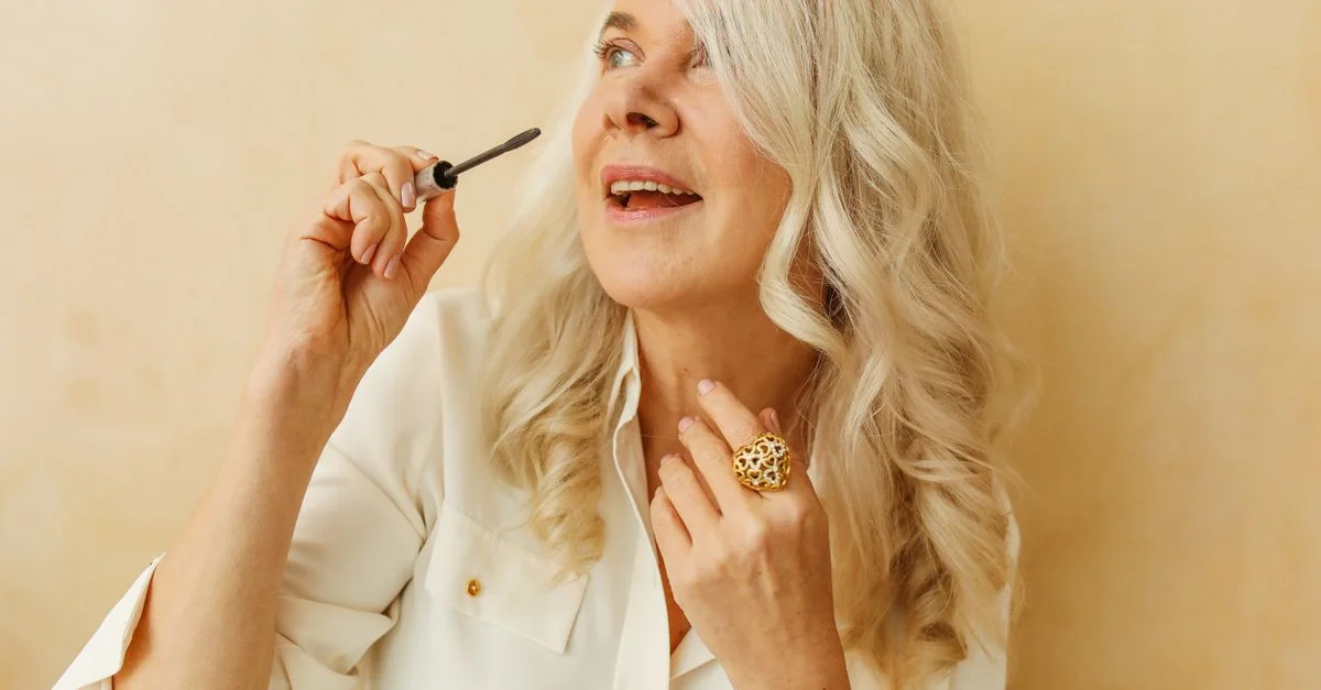 Femme senior joyeuse appliquant du mascara avec un sourire dans un cadre lumineux et stylé.