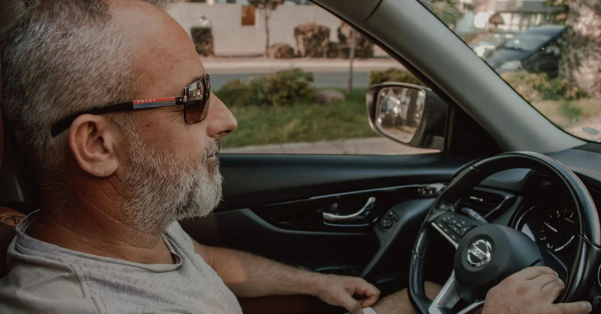 Homme d'âge moyen conduisant une voiture, portant des lunettes de soleil, dégageant une ambiance détendue et confiante.